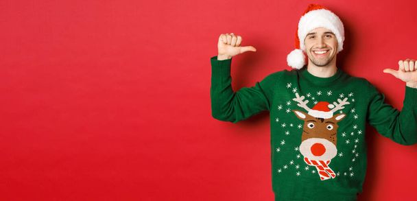 Pojęcie ferii zimowych, Bożego Narodzenia i stylu życia. Szczęśliwy atrakcyjny facet w czapce i swetrze Świętego Mikołaja, wskazujący na siebie z zadowoloną twarzą, stojący nad czerwonym tłem - Zdjęcie, obraz