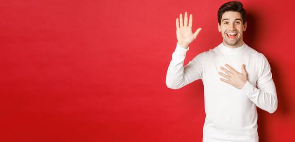 Portrait of honest smiling man in white sweater, making a promise, swearing to tell truth, standing against red background - Foto, afbeelding