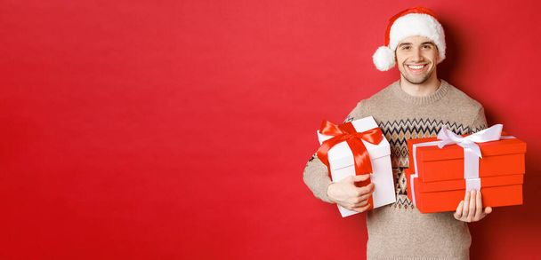 Concept of winter holidays, new year and celebration. Portrait of handsome man in santa hat and sweater, holding boxes with christmas presents and smiling, prepared gifts, red background - Φωτογραφία, εικόνα