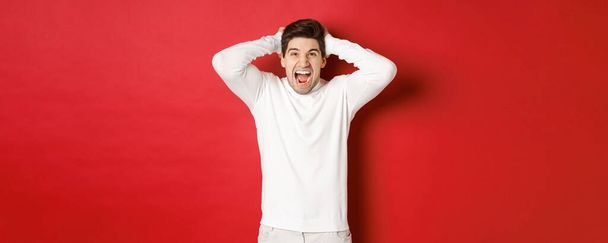 Portrait of frustrated man in white sweater, shouting in anger, holding hands on head and grimacing, feeling distressed, standing over red background - Foto, Imagen