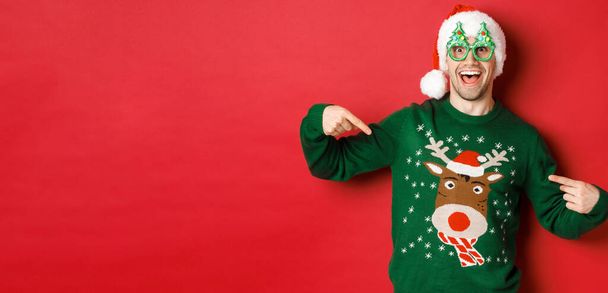 Bild eines glücklichen Mannes mit Partybrille und Weihnachtsmütze, der auf seinen Weihnachtspullover zeigt und lächelt, vor rotem Hintergrund stehend - Foto, Bild