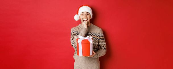 Image of handsome guy in santa hat and christmas sweater, excited to open xmas gift, looking amazed and holding present in one hand, standing over red background - Фото, зображення
