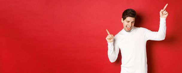Portrait of handsome man enjoying new year party, dancing and having fun, pointing fingers up, standing over red background - Foto, Bild