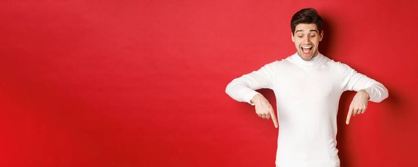 Portrait of happy good-looking man, pointing fingers down and looking at advertisement with excited smile, standing over red background - Foto, imagen