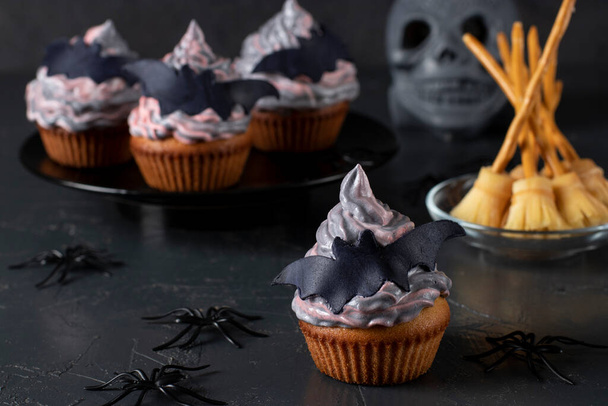 Cupcakes con crema de colores decorados con murciélagos de masilla y escobas de bruja hechas de queso y paja para la fiesta de Halloween, fondo oscuro - Foto, Imagen