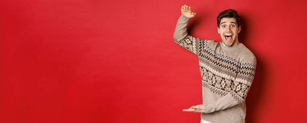 Portrait of excited and happy handsome man in christmas sweater, showing something big, holding large gift for holidays, standing over red background - Photo, Image