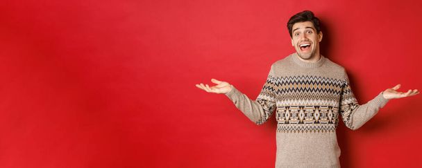 Портрет щасливого і здивованого, красивого кавказького хлопця, одягненого в різдвяний светр, розсунутий руками вбік і виглядає безглуздо, стоїть над червоним тлом
 - Фото, зображення
