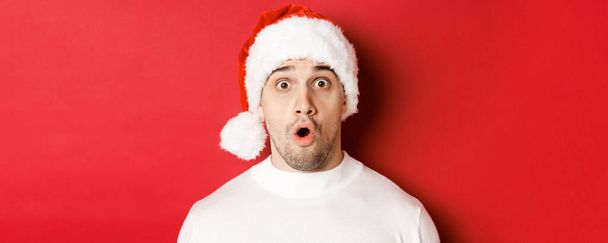 Close-up van aantrekkelijke verraste man in santa hoed, zeggen wow en kijken naar iets interessants, staan tegen rode achtergrond - Foto, afbeelding