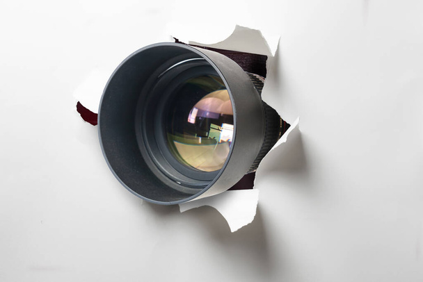 Koncepcja paparazzi lub ukrytej kamery, obiektyw aparatu wychodzi przez otwór w białej ścianie papieru - Zdjęcie, obraz