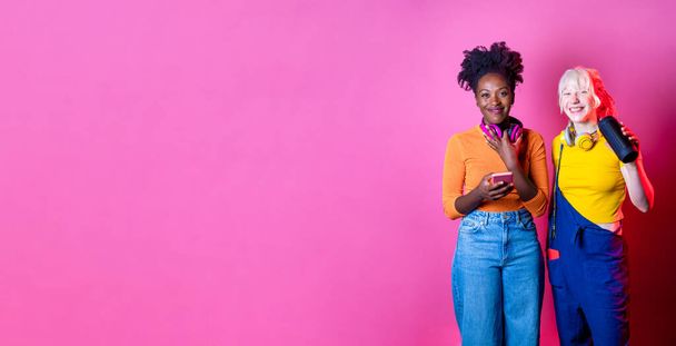 Jóvenes diversas mujeres negras y caucásicas altavoz inalámbrico bluetooth y teléfono inteligente escuchar música lista de reproducción fondo de publicidad aislada - Foto, Imagen
