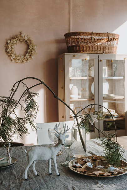 Esthetische zonlicht schaduw reflecties op neutrale stoffige roze muur. Lichte zonnige elegante woon-of eetkamer interieur met dressoir, kerstkrans, dennenboom boeket op linnen tafelkleed - Foto, afbeelding