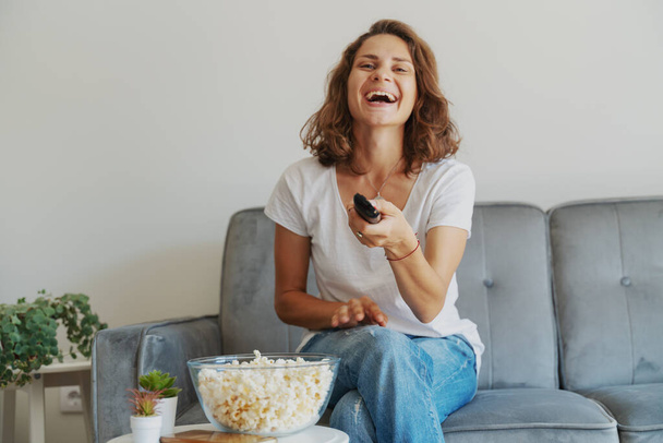 Beautiful smiling happy young woman sitting at home with a tv remote control in her hands and a bowl of popcorn. - Фото, зображення