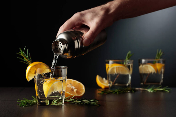 Ο μπάρμαν ρίχνει ένα κοκτέιλ από ένα σέικερ σε ένα ποτήρι. Κοκτέιλ Τζιν και Τονωτικό με λεμόνι και δενδρολίβανο σε μαύρο τραπέζι. - Φωτογραφία, εικόνα