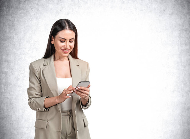 Attractive young smiling businesswoman in formal olive green suit is holding smartphone, dreaming about pleasant things. Woman in business concept, concrete wall on background - Photo, image