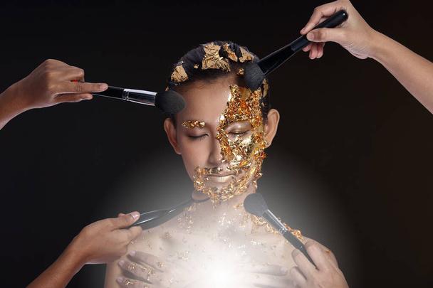 Gold Leaf High fashion style on Asian Woman Face dark mystery look, Golden Mask Facial spa Therapy anti-aging, studio lighting black smoke background, copy space, many hands make up brushes on head - Photo, Image