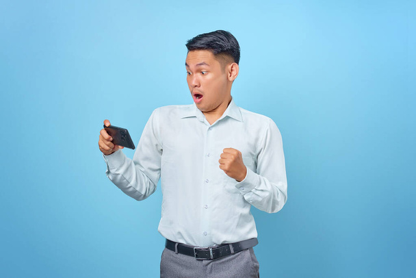 Shocked young handsome businessman looking at smartphone on blue background - 写真・画像