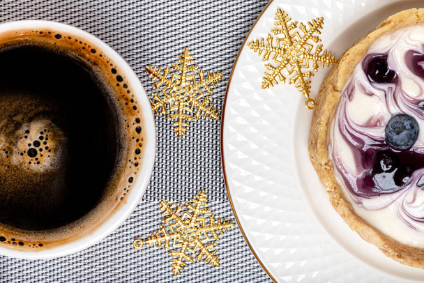 Cerrado de un pedazo de pastel de queso de arándano, una taza de café blace y adornos de Navidad de copo de nieve de oro. - Foto, Imagen