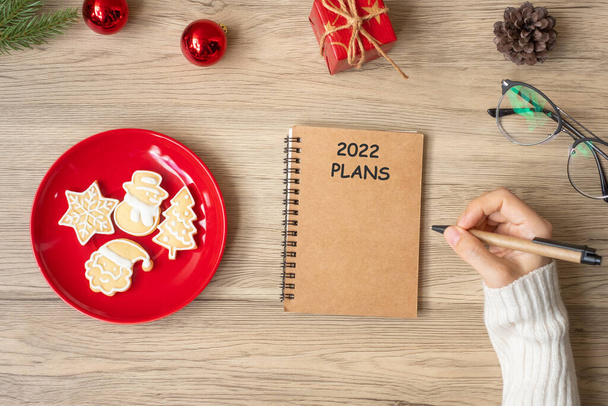 Vrouw handschrift 2022 PLANS op notebook met kerstkoekjes op tafel. Kerstmis, Gelukkig Nieuwjaar, Doelen, Resolutie, Te doen lijst, en Strategie concept - Foto, afbeelding