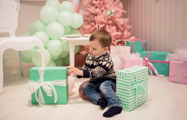маленький мальчик сидит на подушке и распаковывает подарки на фоне рождественской елки - Фото, изображение