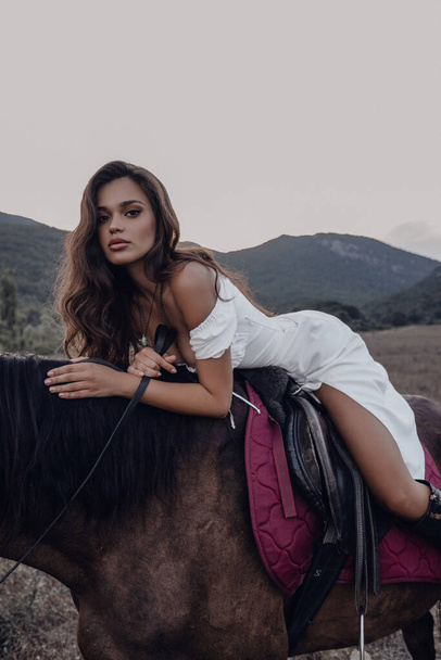fashion outdoor photo of beautiful woman with dark hair in elegant dress posing with horses in the steppe - Photo, image