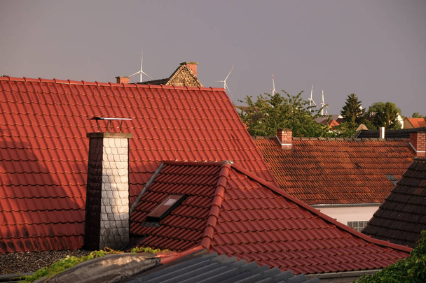 Κόκκινες κεραμοσκεπές γερμανικού χωριού στο φόντο του σταθμού αιολικής ενέργειας. Βραδινή χρυσή ώρα μετά τη βροχή. - Φωτογραφία, εικόνα