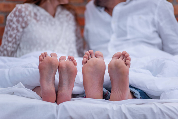 From under the white she covered two pairs of feet of couple of millennials in love. The newlyweds are having fun, wrapped in blanket in the morning on the roof of the open terrace. Lovers spend time - Photo, Image