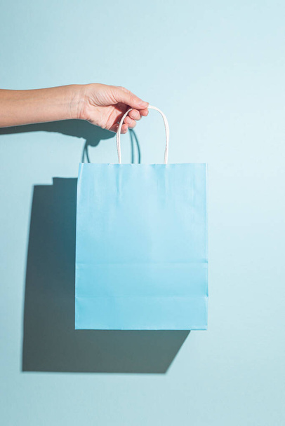 Hand holds a paper shopping bag on a blue background. Hard shadows. - Foto, afbeelding