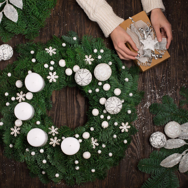 Rustieke kerst sparren krans en handen houden ambachtelijke geschenkdoos op houten tafel, bovenaanzicht. Moderne kerstkrans met kaarsen, takken en sneeuwvlokken op donkere achtergrond. Wintervakantie adventstijd. - Foto, afbeelding