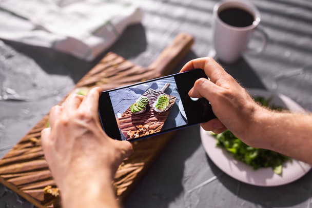 テーブルの上に寝そべっている2つの美しい健康的なサワークリームとアボカドのサンドイッチのスマートフォンで写真を撮る。ソーシャルメディアと食のコンセプト - 写真・画像