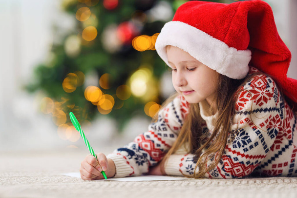 a small child with a Santa hat and New Year's pajamas, lying on the floor, writes a letter with wishes to Santa Claus. Christmas order. removed from the floor level. Merry Christmas and happy holidays - Photo, Image