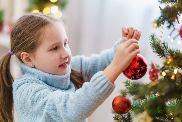 Мила маленька дівчинка прикрашає ялинку вдома. Веселого Різдва. Дитина в синьому жакеті висить на дереві іграшку.. - Фото, зображення