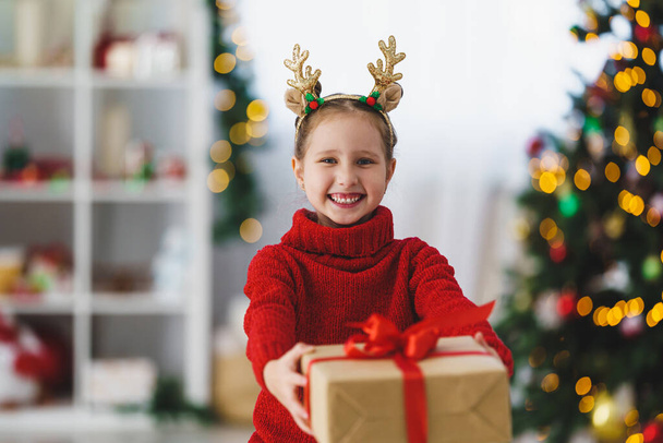 cute cheerful little girl in a red sweater with a box tied with a red ribbon. he holds a gift in his hands, holding it out in front of him. Christmas tree in the light of garlands in the background. - 写真・画像