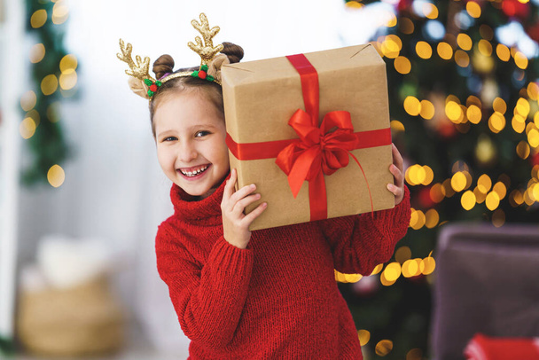 sweet, cheerful, mischievous little girl with a box in her hands, tied with a red ribbon. child is smiling and holding a gift in his hands. Christmas tree in the light of garlands in the background. - Foto, Bild