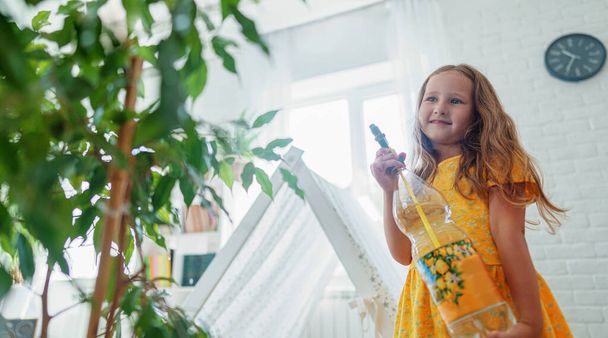 little caring girl sprays water from a spray gun on indoor plants.A child takes care of flowers, sprinkles water from a bottle on a palm tree plant. The baby helps with cleaning the house. - Foto, Imagen