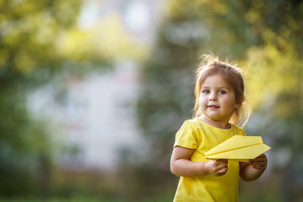 portrait of cute little girl in yellow T-shirt stands holding paper airplane in her hands. child plays role of pilot launching a paper airplane into the air. kid. Children's active play in fresh air. - Фото, изображение