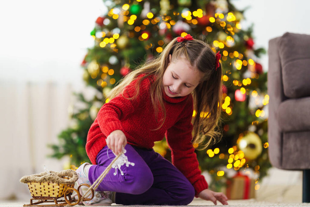 A happy little girl is playing with a toy in a room decorated for Christmas. Family Christmas morning in a decorated living room with children's gifts, a Christmas tree. - Fotoğraf, Görsel