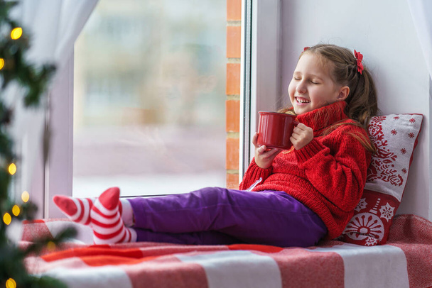 A cute little girl is sitting by the window with a mug of hot drink. Decorated Christmas tree. a child in warm clothes with a blanket and a pillow by the window. Merry Christmas and happy holidays. - Foto, afbeelding