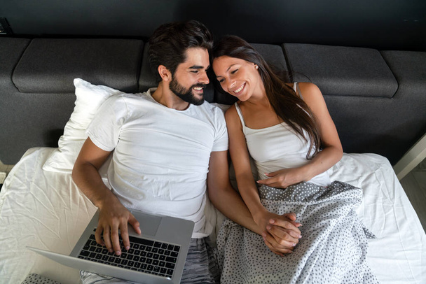 Happy young man and woman is having fun in bed together. Couple technology concept. - Foto, afbeelding