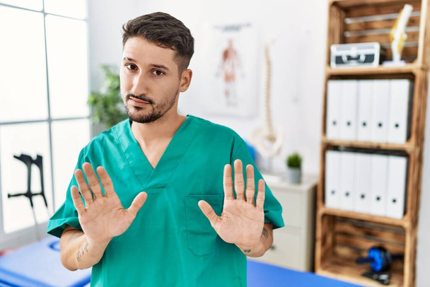 Young physiotherapist man working at pain recovery clinic moving away hands palms showing refusal and denial with afraid and disgusting expression. stop and forbidden.  - Photo, Image