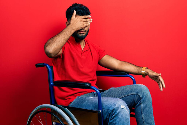Hombre árabe con barba sentado en silla de ruedas cubriendo los ojos con la mano, luciendo serio y triste. concepto de ocultamiento y rechazo sin visión  - Foto, imagen