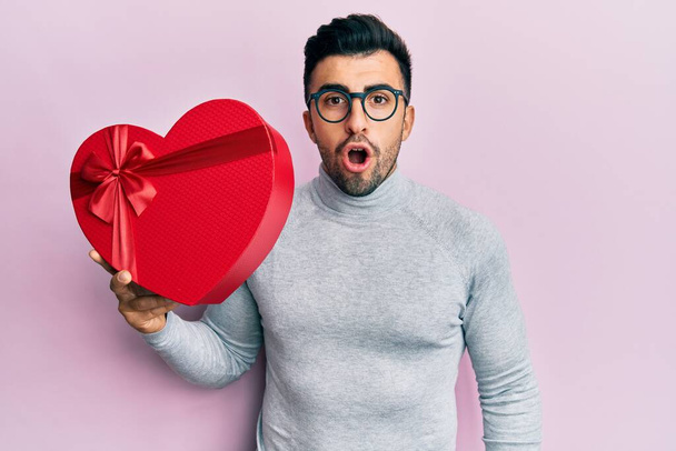 Joven hombre hispano sosteniendo regalo de San Valentín asustado y sorprendido con la boca abierta para sorpresa, cara de incredulidad  - Foto, Imagen