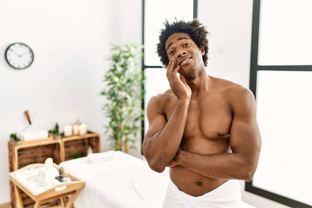 Młody, afrykański mężczyzna bez koszulki, noszący ręcznik stojący w centrum piękności, myślący, że wygląda na zmęczonego i znudzonego problemami z depresją z skrzyżowanymi ramionami..  - Zdjęcie, obraz