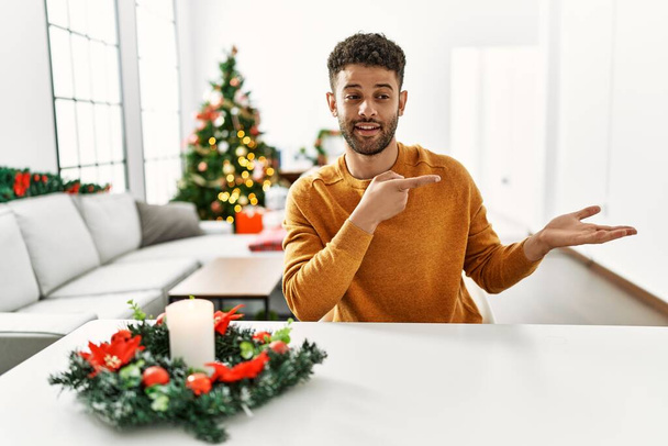 Arabischer junger Mann sitzt erstaunt auf dem Tisch am Weihnachtsbaum und lächelt in die Kamera, während er mit der Hand präsentiert und mit dem Finger zeigt.  - Foto, Bild