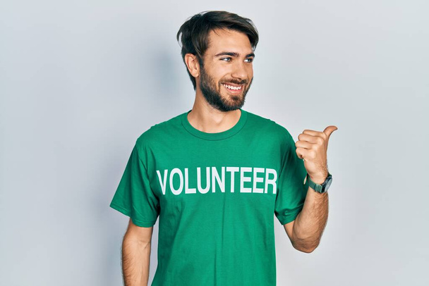 Молодой латиноамериканец в футболке добровольца улыбается со счастливым лицом и указывает на сторону с большим пальцем вверх.  - Фото, изображение