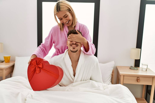 Woman surprising her boyfriend with valentine gift box sitting on the bed at home. - Photo, Image