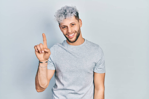 Joven hombre hispano con el pelo teñido moderno con camiseta gris casual mostrando y señalando con el dedo número uno mientras sonríe confiado y feliz.  - Foto, Imagen