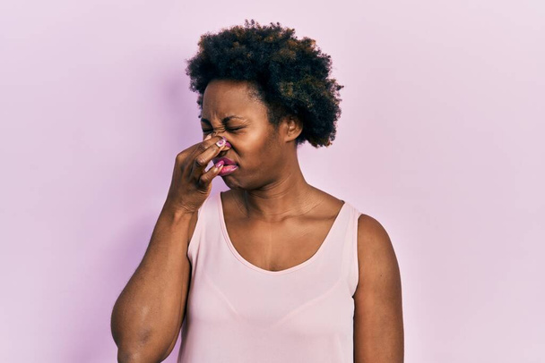 Mujer afroamericana joven que usa una camiseta informal sin mangas oliendo algo apestoso y asqueroso, olor intolerable, conteniendo la respiración con los dedos en la nariz. mal olor  - Foto, Imagen