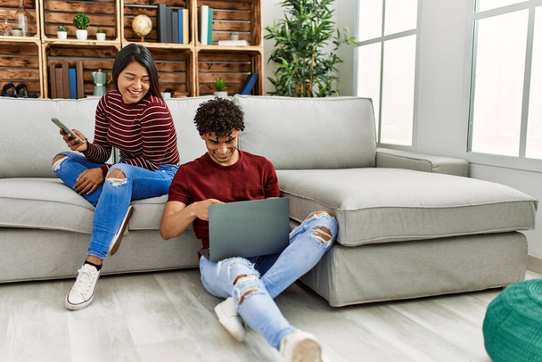 Молода латинська пара посміхається щасливий, використовуючи ноутбук і смартфон вдома
. - Фото, зображення