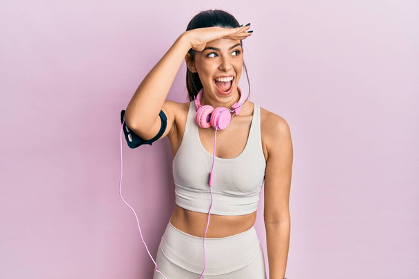Young hispanic woman wearing gym clothes and using headphones very happy and smiling looking far away with hand over head. searching concept.  - Photo, Image