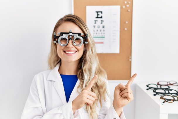 Fiatal, gyönyörű optikus nő, szemüveget visel a klinikán, mosolyog és a kamerába néz, két kézzel és ujjal oldalra mutatva..  - Fotó, kép